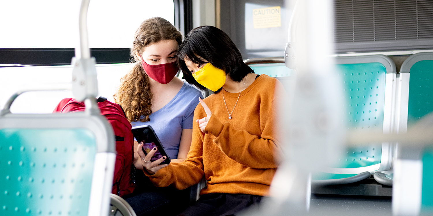 Two woman with masks, sitting on the bus, watching something on a smartphone