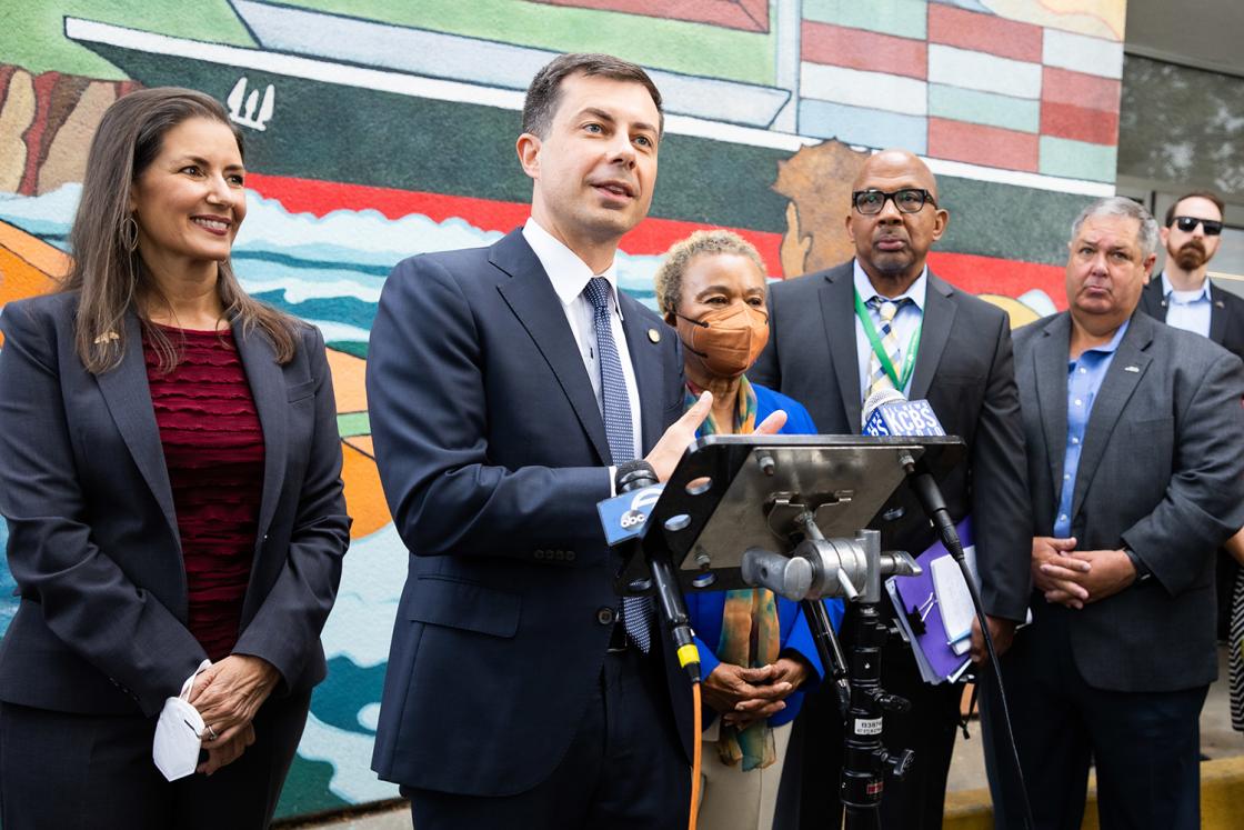 Secretary Pete Buttegieg speaking to media, Rep. Barbara Lee, Oakland Mayor Libby Shaff, and AC Transit GM Michael Hursh standing behind him