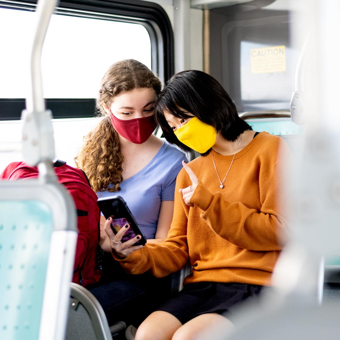 Two girls with masks on, sitting on the bus, looking at a phone screen